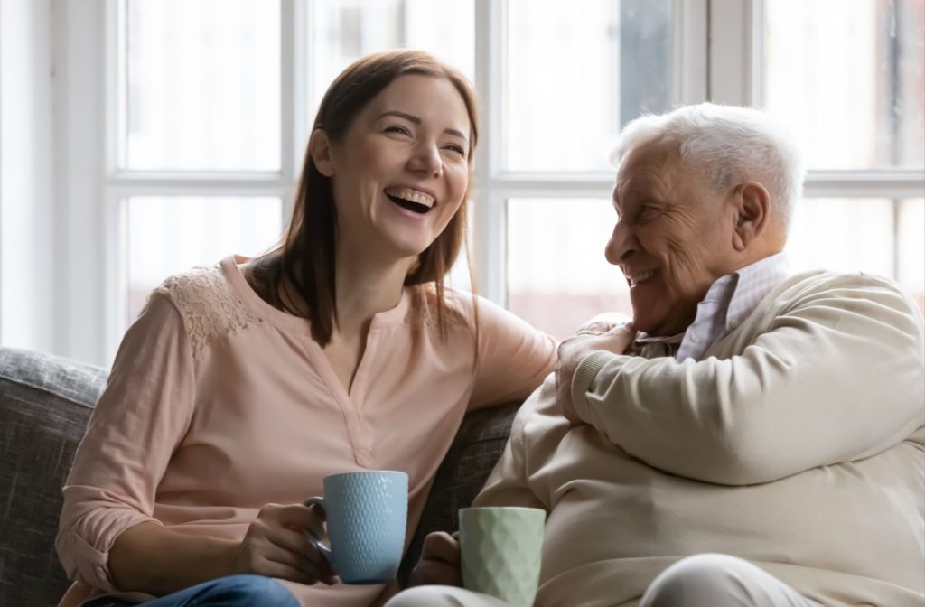 Someone visits their parent in their memory care home, the two laughing over coffee.
