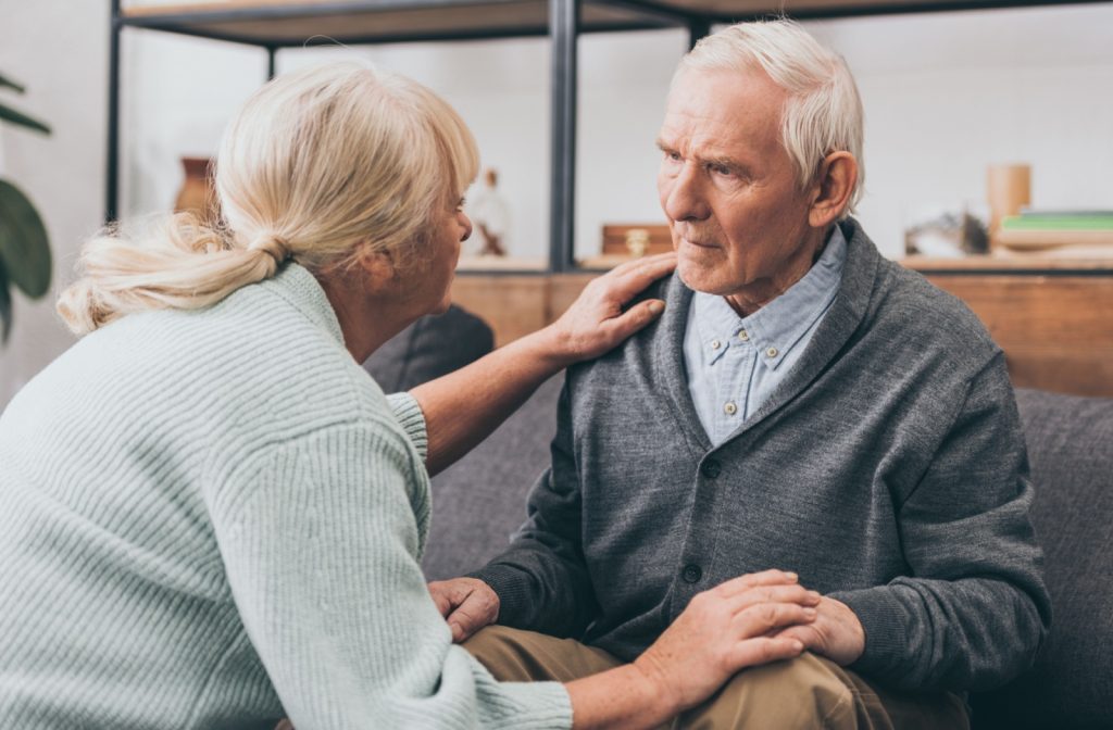 An older adult kneels to check on their spouse on the couch at home and rubs their shoulder to reassure them about their recent memory loss