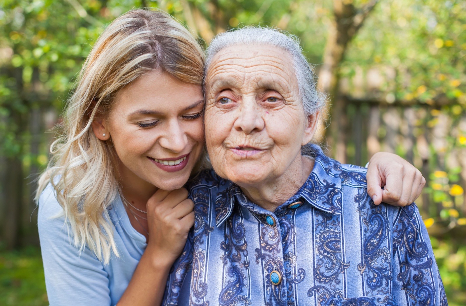 A smiling adult child embraces their senior parent while sitting outside on a sunny day.
