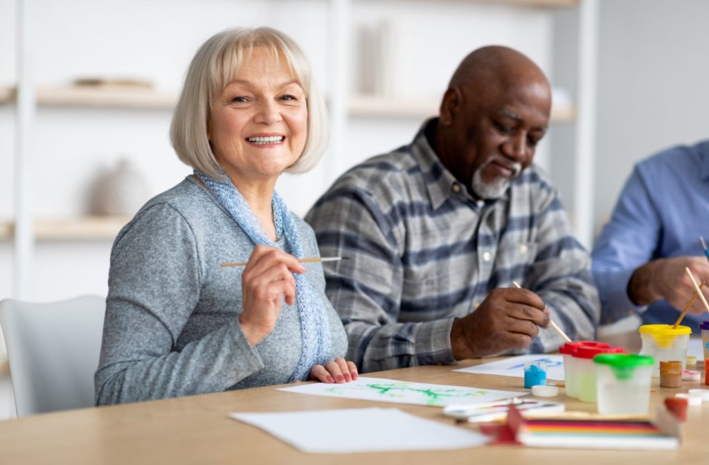 A senior smiles at the camera as they attend a painting class with friends.